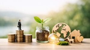 Une figurine d’homme d’affaires debout sur des piles de pièces de monnaie, à côté d'une jeune plante poussant dans un bocal en verre. À droite, une ampoule allumée est posée près de silhouettes en bois représentant le globe terrestre, un cerf, une maison et un cœur, sur fond vert naturel flouté.